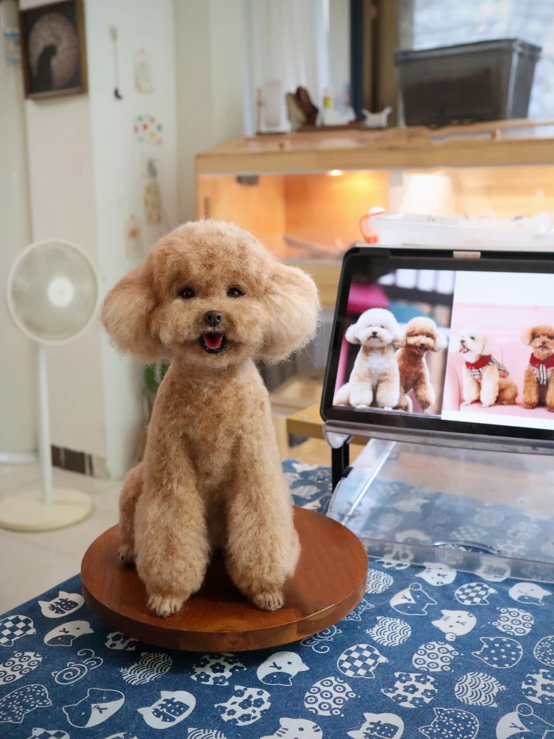 A meticulously groomed Teddy Bear dog sits on a table covered with a blue cat-patterned tablecloth. It has fluffy brown fur and a cute expression. In the background, a tablet displays images of other similarly styled pets.