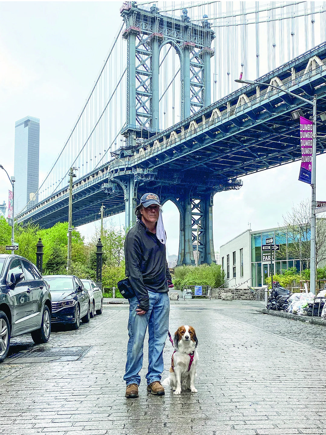 A man leads a brown and white Papillon dog while standing beneath a majestic bridge, with iconic city architecture and a street in the background. The dog sits curiously, and the man looks straight ahead, depicting a scene of urban life with a pet.