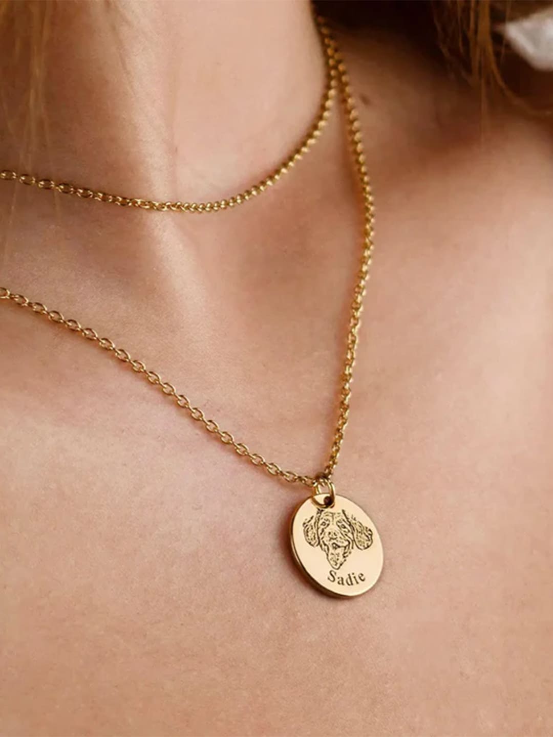 A delicate golden necklace is worn around a woman's neck, with a pendant engraved with a pet dog's head and the name "Sadie". This is a personalized pet memorial jewelry piece, expressing love and remembrance for a beloved pet.