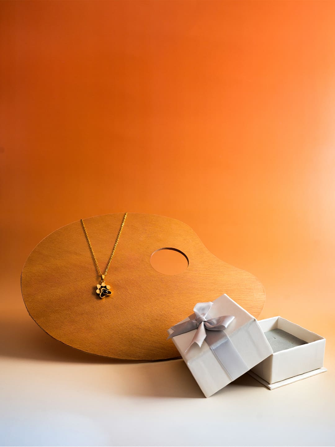 Gold flower pendant necklace on artist's palette with gift box (silver ribbon). Orange background; stylish gift.